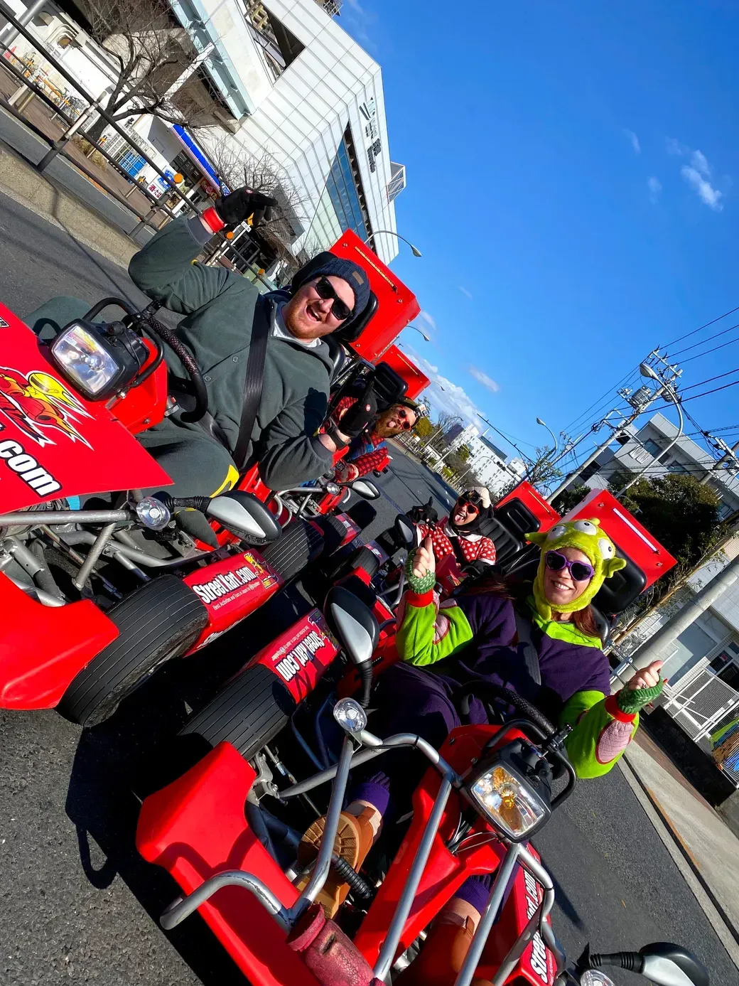 Four friends in colorful outfits pose on bright red pedal go‑karts on a city street, smiling and giving thumbs up under a blue sky.