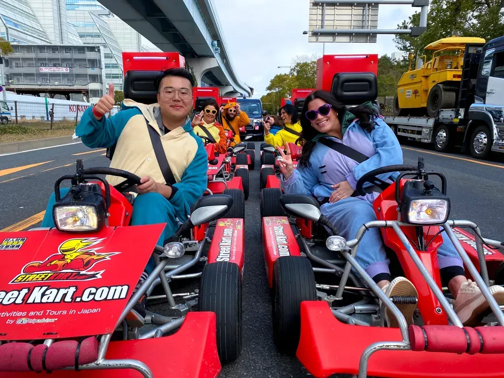 Group of friends in red go‑karts on a city street, smiling and giving thumbs up to the camera.