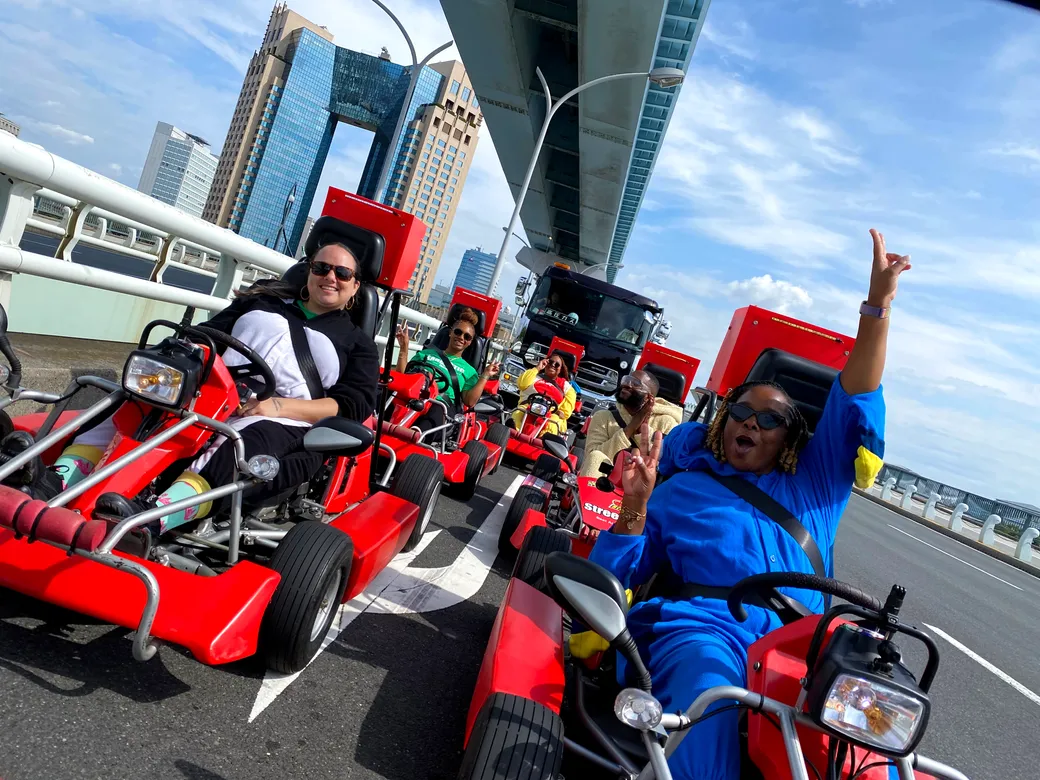 Group of people riding red pedal go-karts on a city bridge with tall buildings in the background; one rider raises an arm in the air.