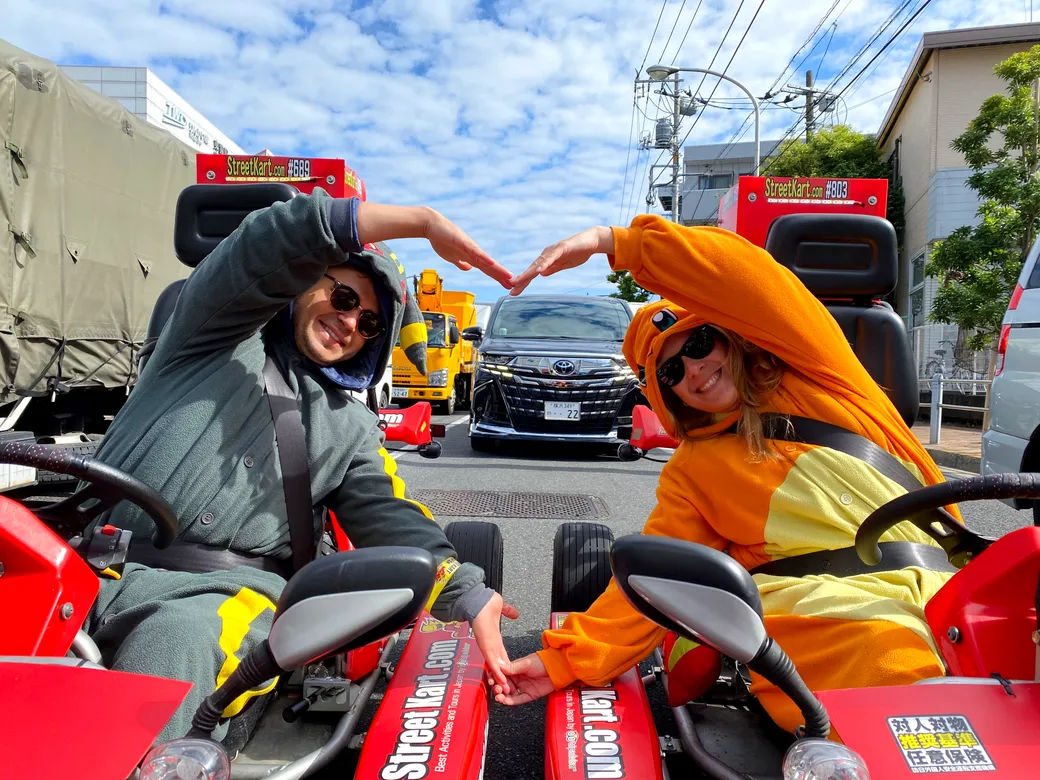Two friends in red go-karts on a city street form a heart shape with their arms and smile at the camera, wearing colorful outfits (gray and orange).